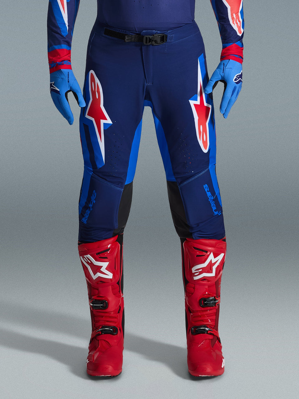 Alpinestars 2026 Supertech Vista Pants, Motocross Pants, Dark Navy, Hot Coral, and Cobalt, front view of rider wearing performance gear with matching blue gloves and red motocross boots, featuring prominent red and white Alpinestars logos on thighs.