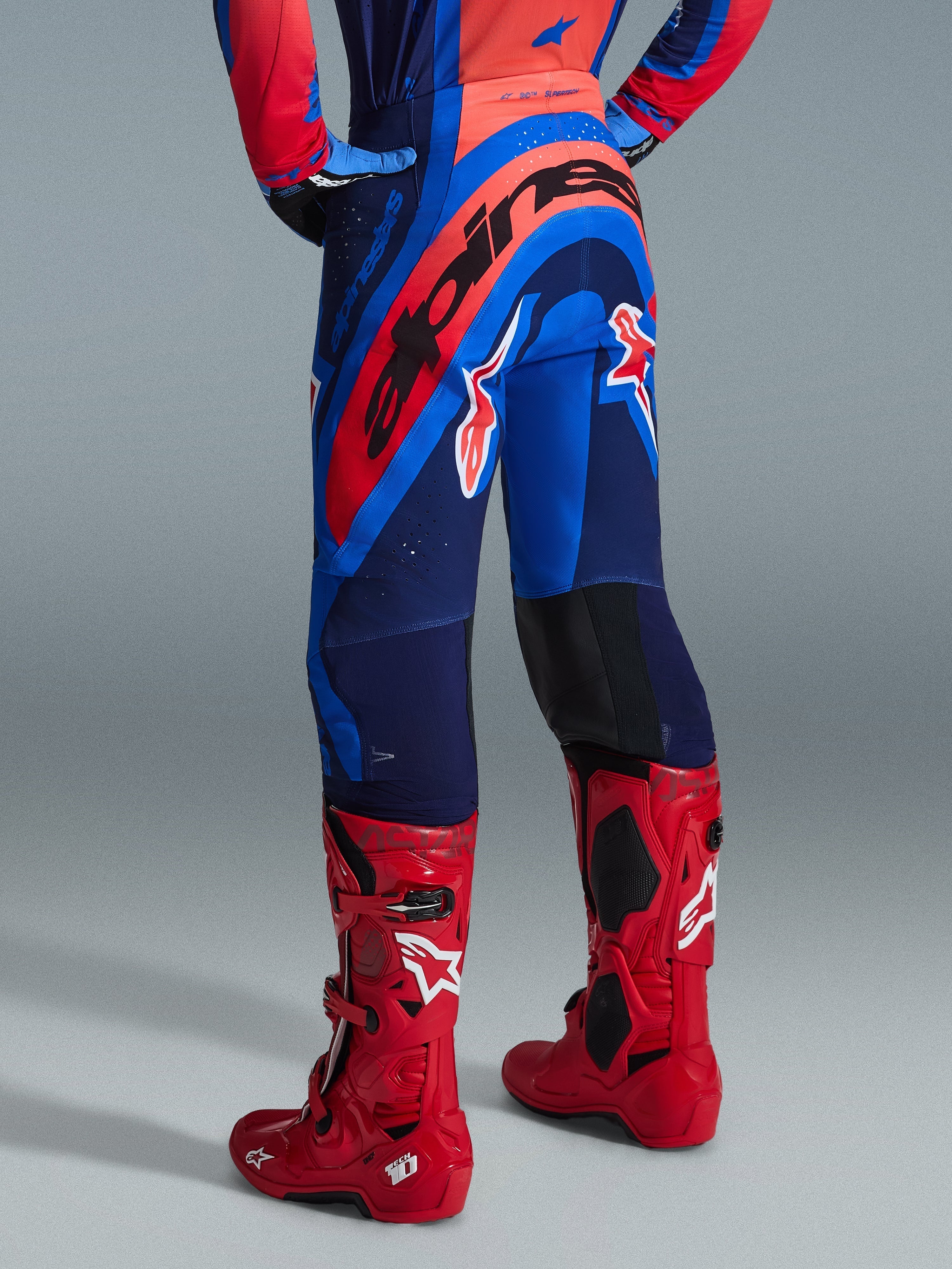 Alpinestars 2026 Supertech Vista byxor, motocrossbyxor, Dark Navy Hot Coral Cobalt, vy bakifrån som visar en framträdande röd Alpinestars-logotyp över sätet, matchade med klarröda motocross-stövlar, lättviktig stretchkonstruktion för professionell racing.