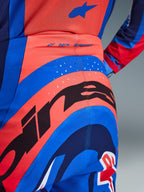 Närbild på Alpinestars 2026 Supertech Vista-byxor, motocrossbyxor i Hot Coral och Cobalt Blue, med en stor svart Alpinestars-grafik på sätet, laserskurna ventilationshål och 'SUPERTECH'-logotyp i midjan bak, tävlingskläder av proffsklass.