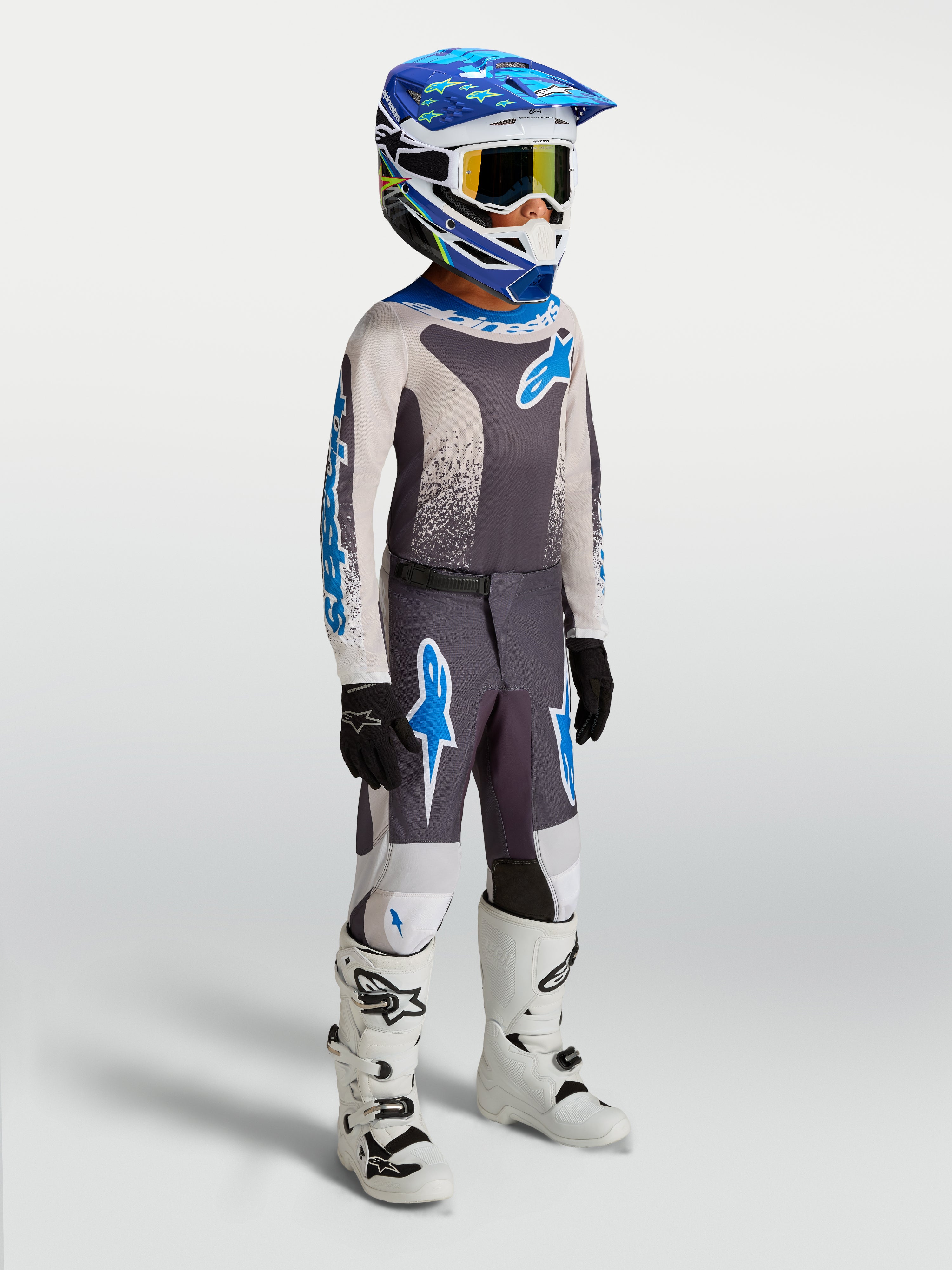 Alpinestars 2027 Ungdom Fluid Drag byxor, motocrossbyxor för ungdomar, gråa och blåa, bärs av modell med matchande tröja och hjälm, slitstark 600D-konstruktion för terrängkörning