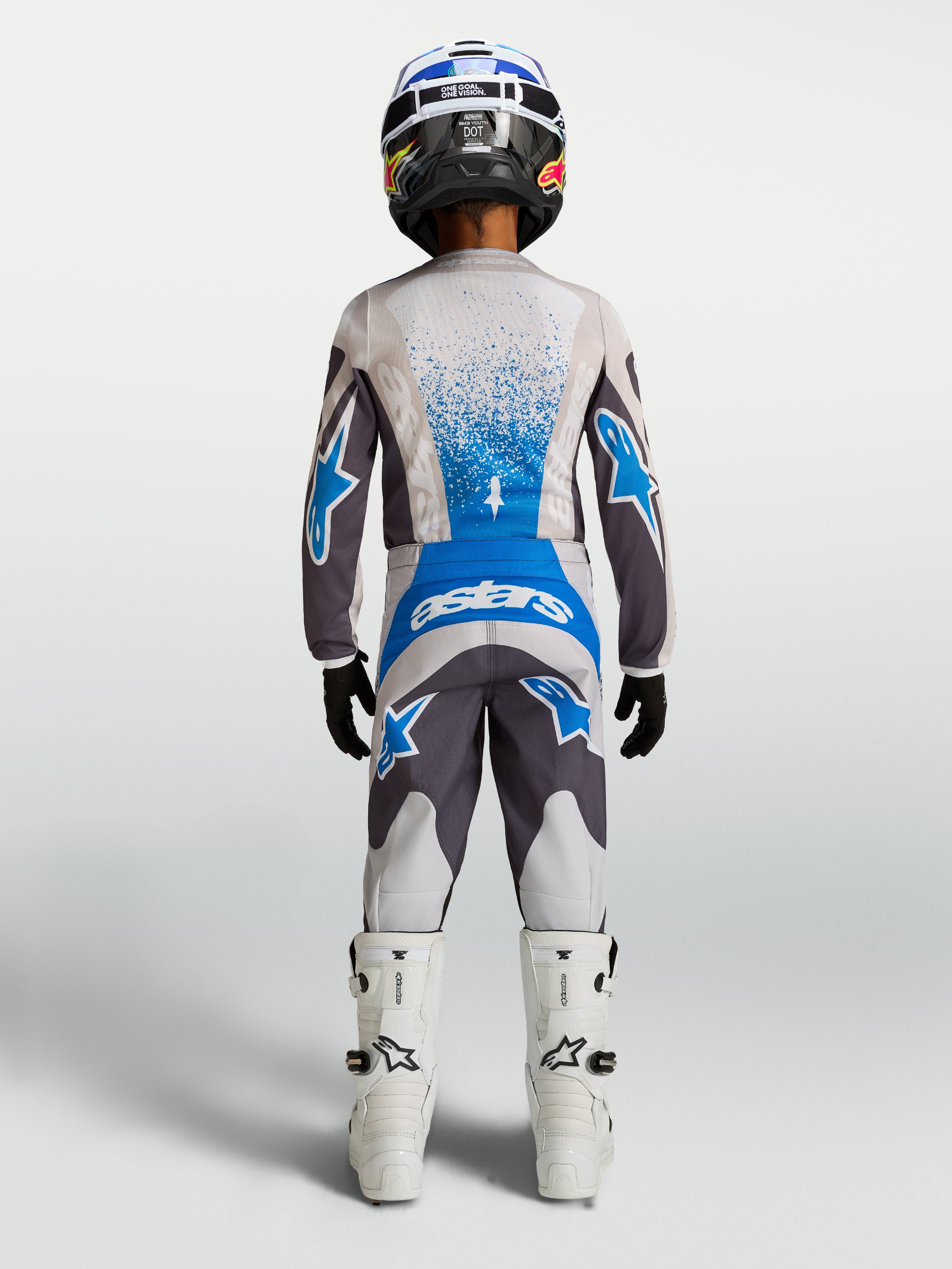 Alpinestars 2027 Ungdom Fluid Drag byxor, motocrossbyxor för ungdom i grått och blått, vy bakifrån bärs med matchande tröja och vita motorcykelstövlar, slitstark off-road-design.