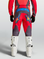 Alpinestars 2027 Fluid Hunt ungdomsbyxor, röda och grå motocrossbyxor för terrängkörning, vy bakifrån med blå grafik, hållbart 600D-material och vita motorcykelstövlar.