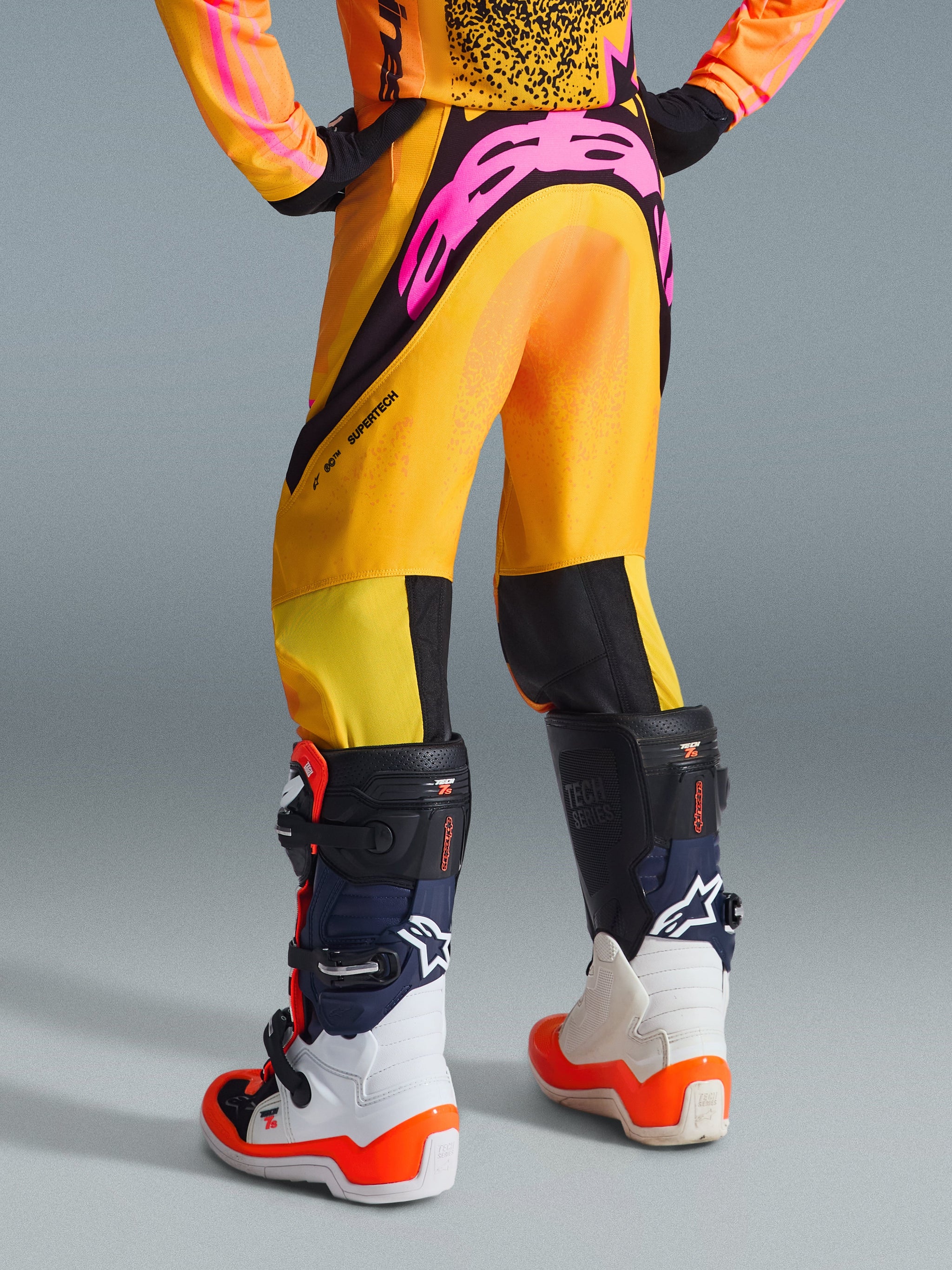 Alpinestars 2026 Youth Supertech Nomur Pants, Motocross Pants, Orange Yellow and Pink, rear view showing large pink logo on seat, paired with white and blue Tech 7S boots, professional racing gear for youth off-road riders