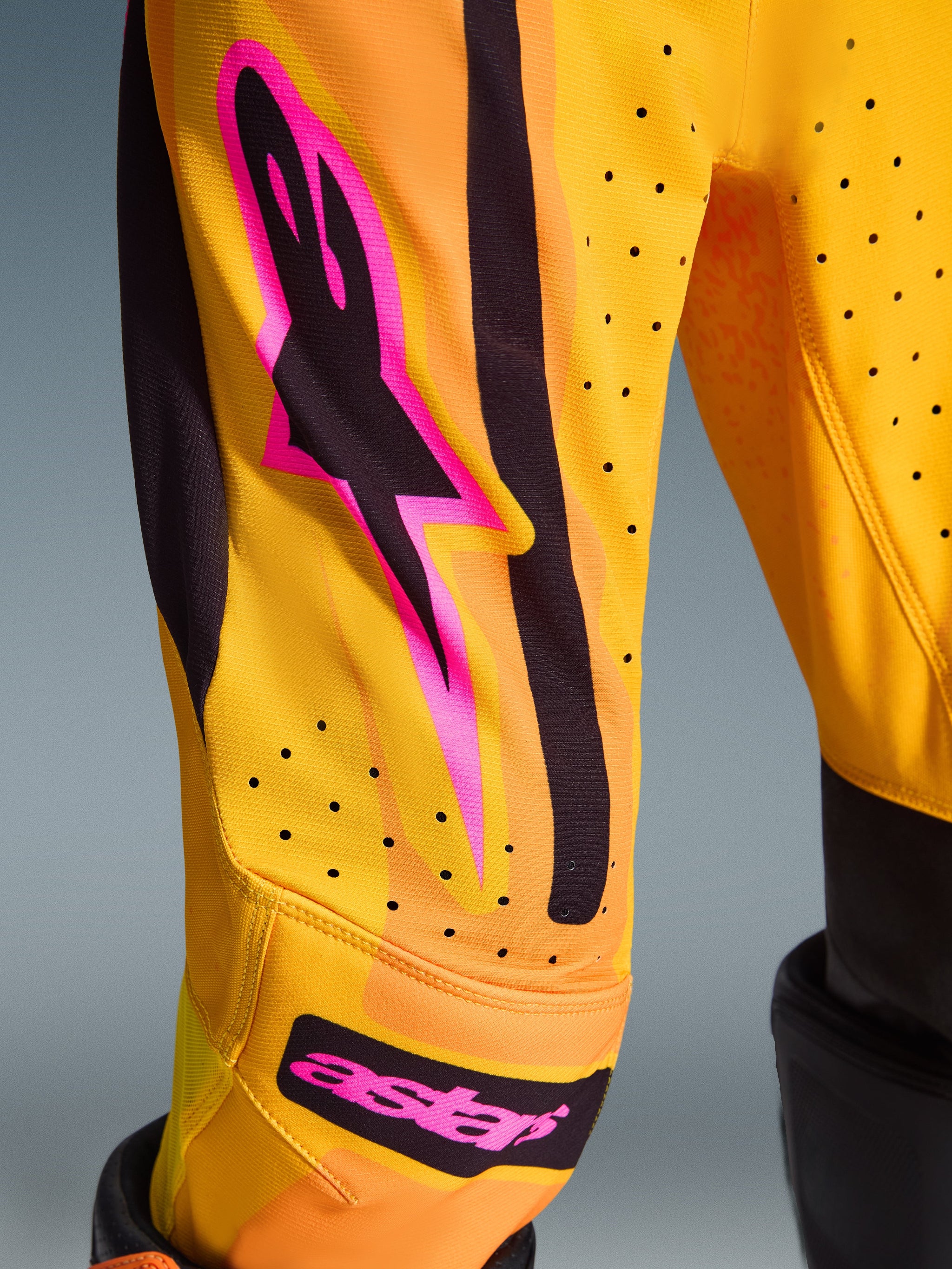 Alpinestars 2026 Youth Supertech Nomur Pants, Motocross Pants, Orange Yellow and Pink, close-up of the leg area featuring pink and black Alpinestars logo, perforated ventilation holes, and vibrant graphics