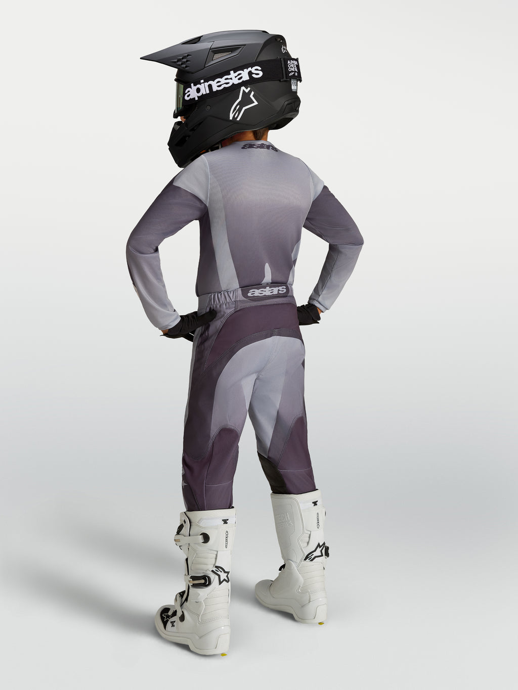 Alpinestars Fluid Amber ungdomsbyxor, grå och mörklila sportiga motocrosskläder, visas med vita motorcykelstövlar.