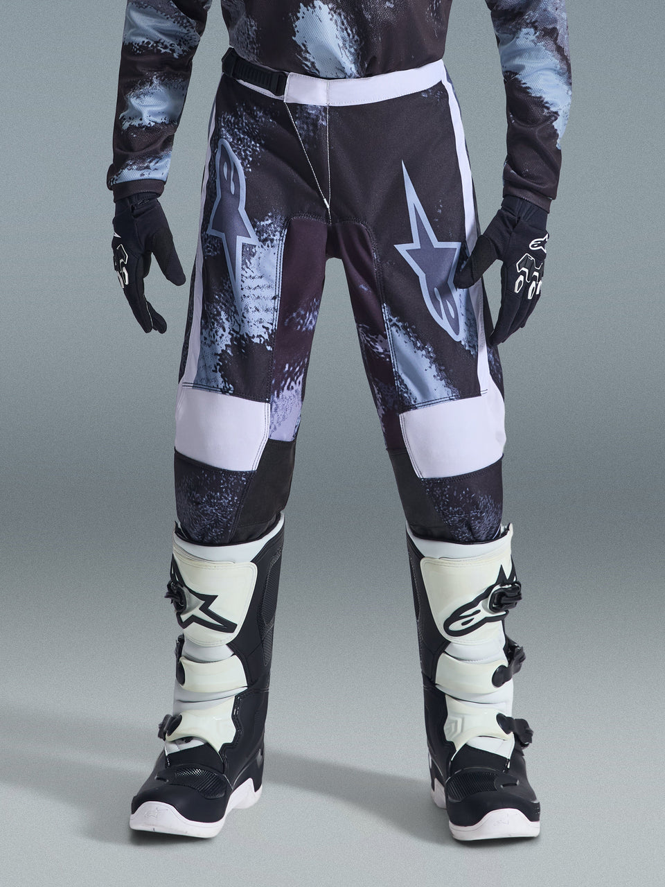 Alpinestars 2026 Youth Fluid Lahnd-byxor, motocrossbyxor för ungdom, Iron Camo med vita detaljer, bärs med svartvita offroad-stövlar och handskar, slitstark design för unga förare.