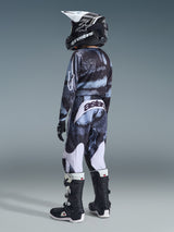 Alpinestars 2026 Youth Fluid Lahnd byxor, motocrossbyxor, Iron Camo-mönster, ungdomsförare sedd bakifrån i matchande camotröja, svarta och vita stövlar och svart hjälm, med vita paneler och 'astars'-logotyp på baksidan, komplett utrustning för terrängkörning