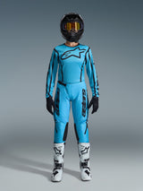 Female motocross rider wearing the 2026 Alpinestars Stella Fluid Apex gear set, bright cyan blue jersey and pants with black accents, black helmet with iridescent goggles, and white boots, full professional motocross outfit