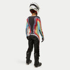 Motocross rider from behind wearing Alpinestars Women Stella 2024 Techstar Pants in Black Multicolor, featuring large white branding on the seat, coordinated with a colorful striped jersey, white helmet, and black Tech 3 boots for off-road racing.