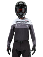 Motocrossförare som bär Alpinestars Pro-Dura Jersey i Black Magnet White, långärmad funktionströja med vita och grå färgblock, svart hjälm och goggles, 'alpinestars'-branding och slogan 'ONE GOAL ONE VISION', offroad-racingkläder