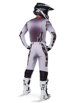 Motocrossförare sedd bakifrån iförd Alpinestars 2025 Supertech Maker-byxor i Silver och Svart, matchande långärmad tröja, svartvit hjälm och vita stövlar med svarta detaljer, framträdande svart logotyp på benen, professionell utrustning för terrängkörning