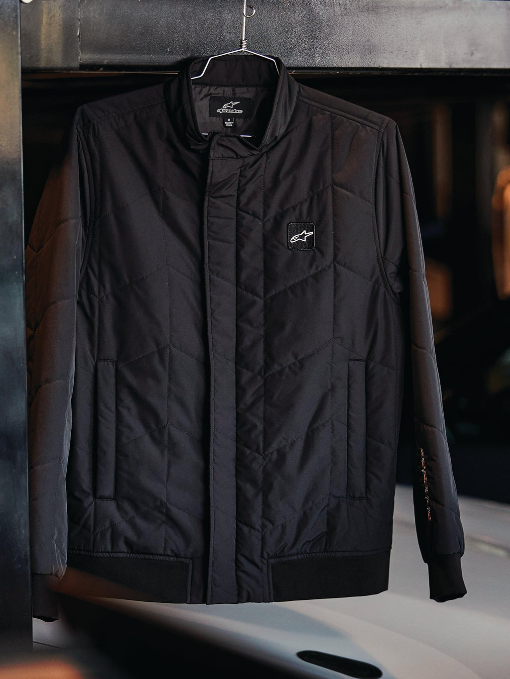 Alpinestars Precedent-jacka, quiltad sportswear-jacka, svart, med vinklat quiltmönster, låg ståkrage med täckt dragkedja, logomärke på bröstet, ribbade muddar och nederkant, motorsport-inspirerad design