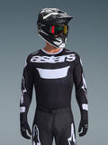 Alpinestars 2026 Racer Air Riway Jersey, svart och vit motocrosströja med 'astars'-profilering, bärs av en förare med matchande svart hjälm, goggles och byxor, med ventilerande meshpaneler för terrängkörning.