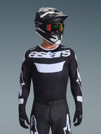 Alpinestars 2026 Racer Air Riway Jersey, svart och vit motocrosströja med 'astars'-profilering, bärs av en förare med matchande svart hjälm, goggles och byxor, med ventilerande meshpaneler för terrängkörning.