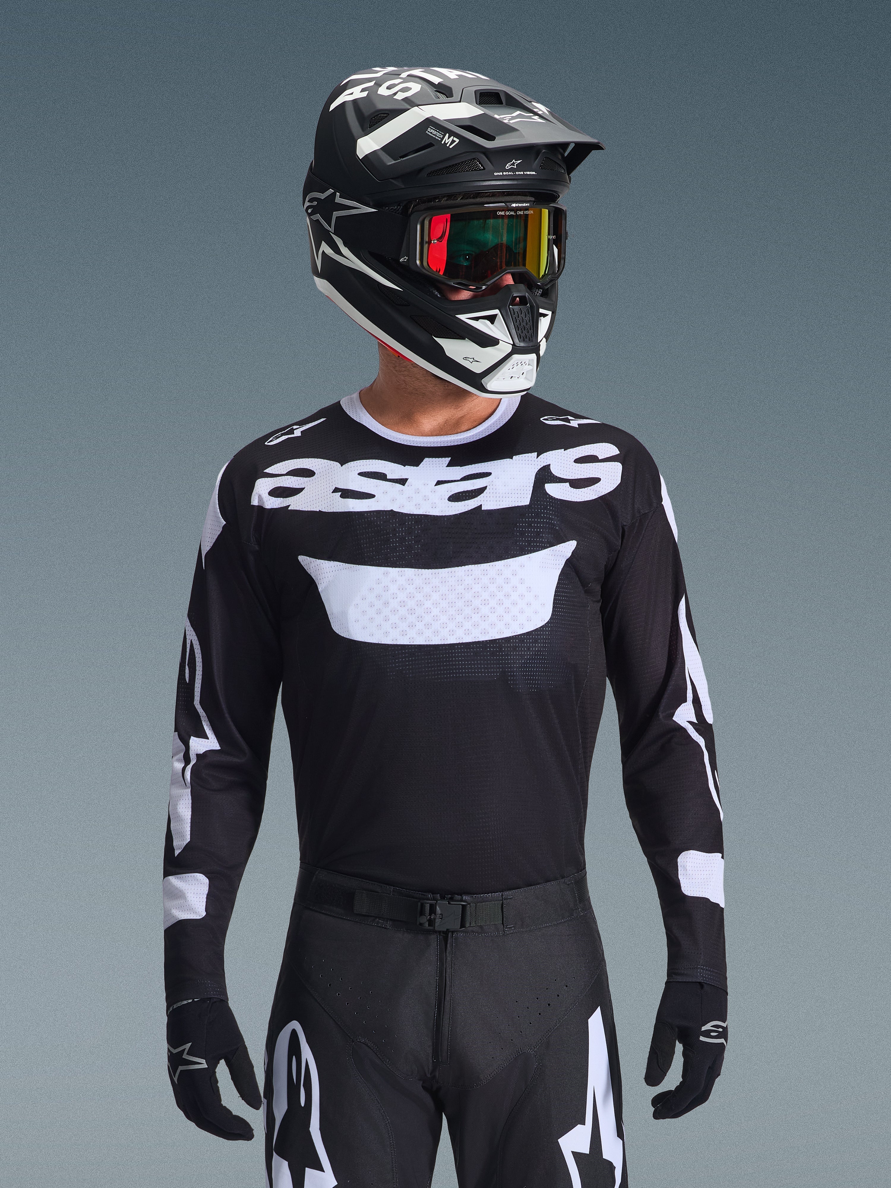 Alpinestars 2026 Racer Air Riway Jersey, svart och vit motocrosströja med 'astars'-profilering, bärs av en förare med matchande svart hjälm, goggles och byxor, med ventilerande meshpaneler för terrängkörning.