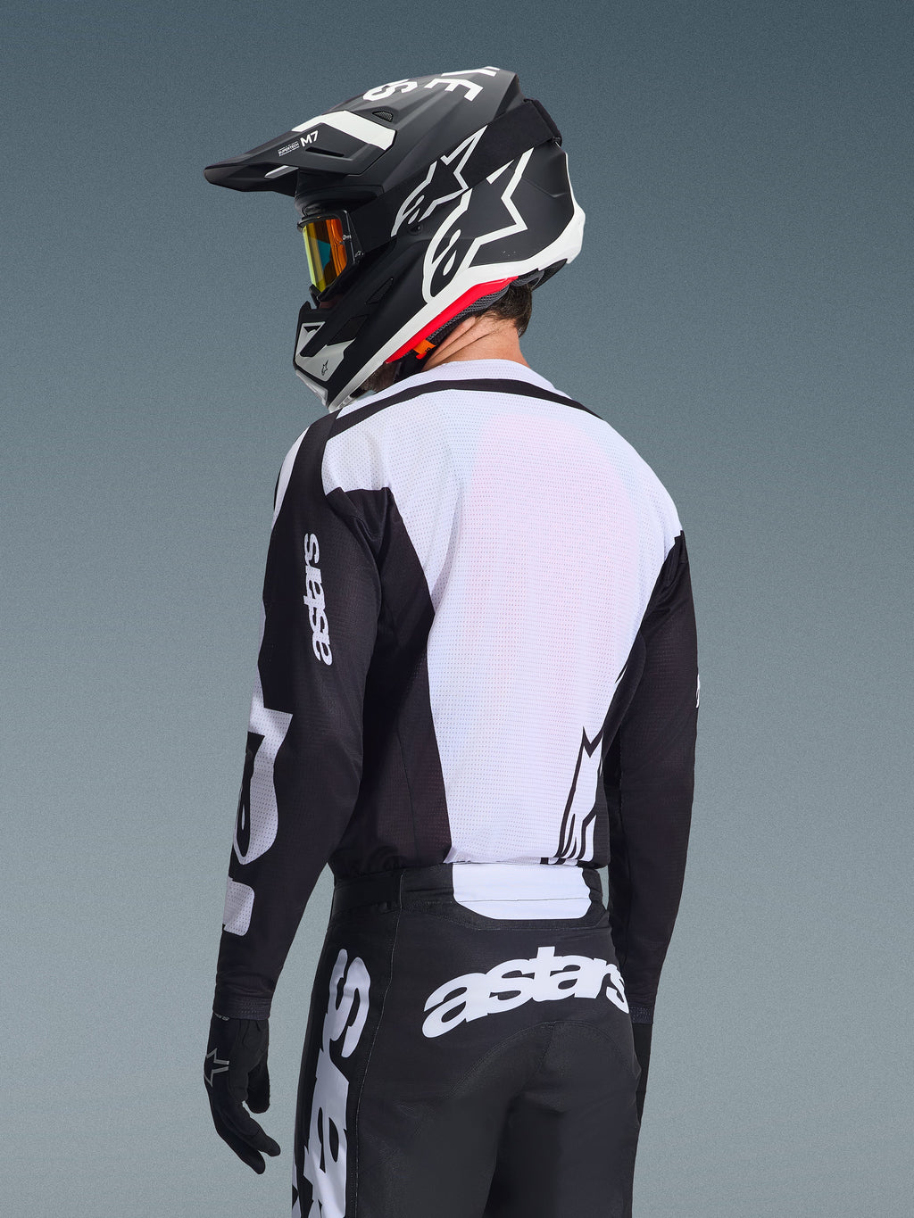 Motocrossförare sedd bakifrån iförd Alpinestars 2026 Racer Air Riway Jersey, svart och vit, med vit ryggpanel i ventilerande mesh och svarta ärmar med 'astars'-logotyp, matchande svarta byxor med vita logotyper, svart hjälm med vita detaljer, professionell offroad-utrustning för prestanda i höga temperaturer.
