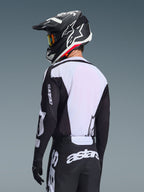 Motocrossförare sedd bakifrån iförd Alpinestars 2026 Racer Air Riway Jersey, svart och vit, med vit ryggpanel i ventilerande mesh och svarta ärmar med 'astars'-logotyp, matchande svarta byxor med vita logotyper, svart hjälm med vita detaljer, professionell offroad-utrustning för prestanda i höga temperaturer.