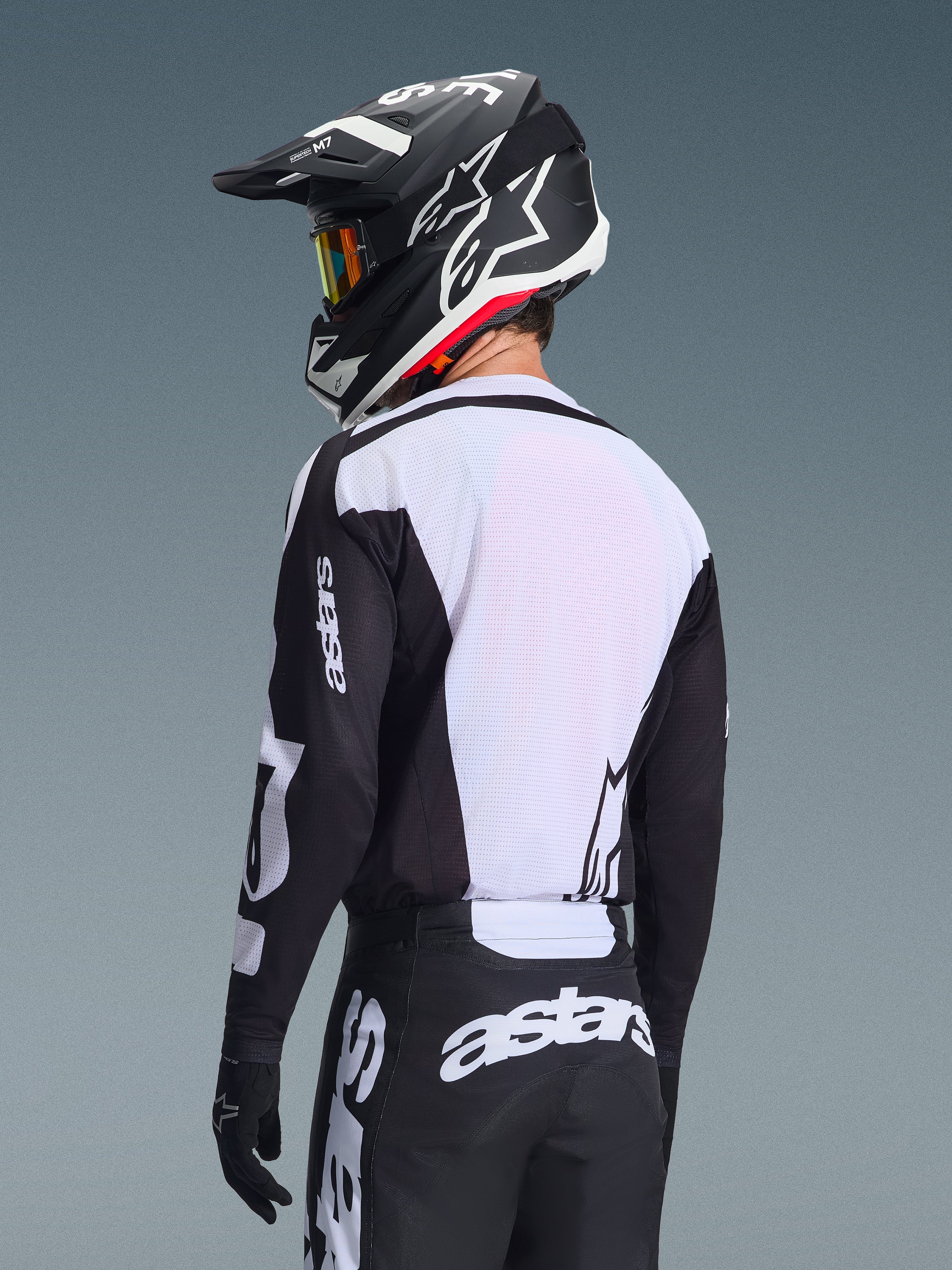 Motocrossförare sedd bakifrån iförd Alpinestars 2026 Racer Air Riway Jersey, svart och vit, med vit ryggpanel i ventilerande mesh och svarta ärmar med 'astars'-logotyp, matchande svarta byxor med vita logotyper, svart hjälm med vita detaljer, professionell offroad-utrustning för prestanda i höga temperaturer.