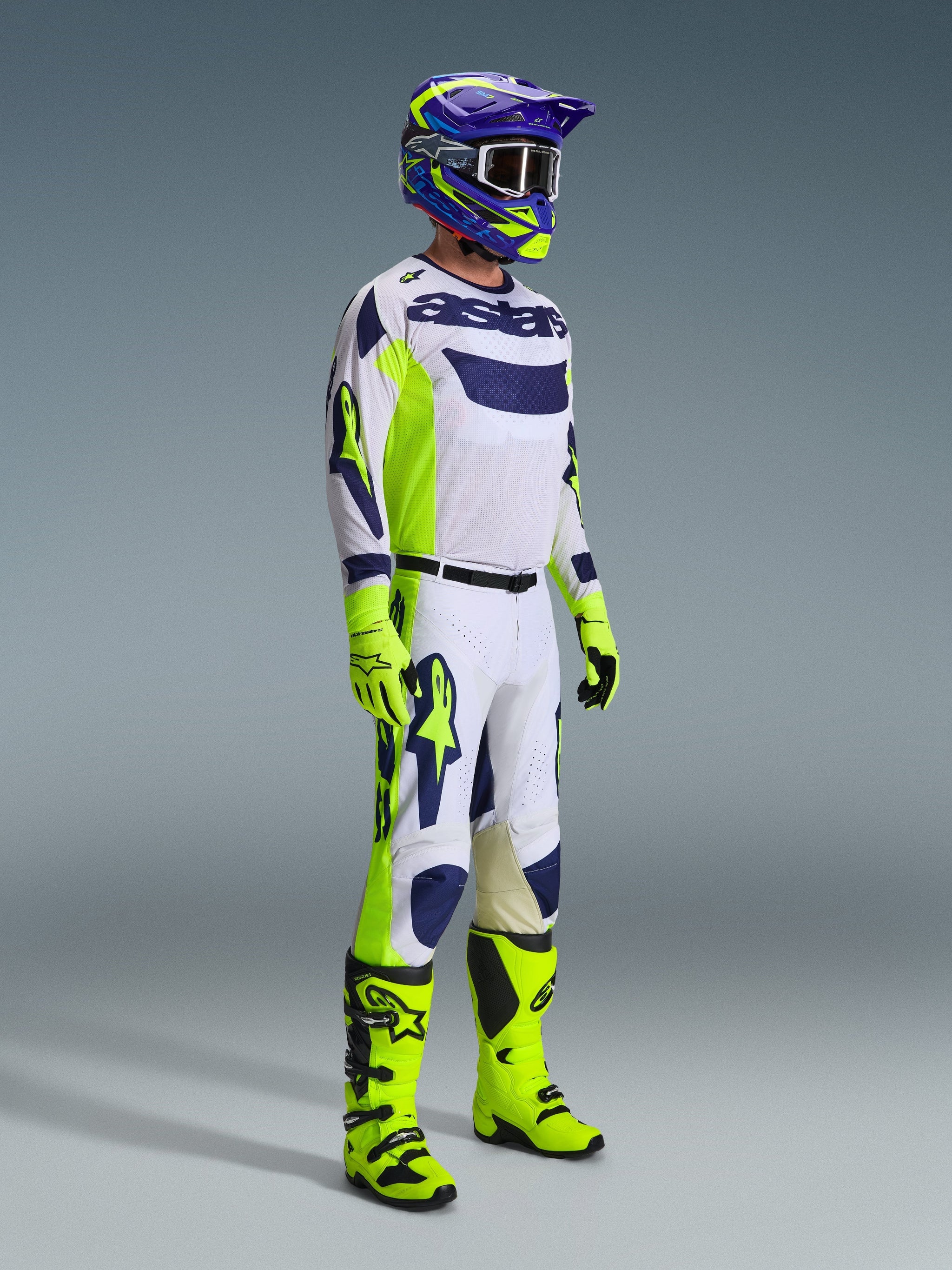 Motocrossförare i komplett racingutrustning, ljusgrå tröja med Yellow Fluo och blå detaljer, matchande vita och gula byxor, neongula stövlar och handskar, lila och gul hjälm med mörka goggles, koordinerad outfit för terrängkörning
