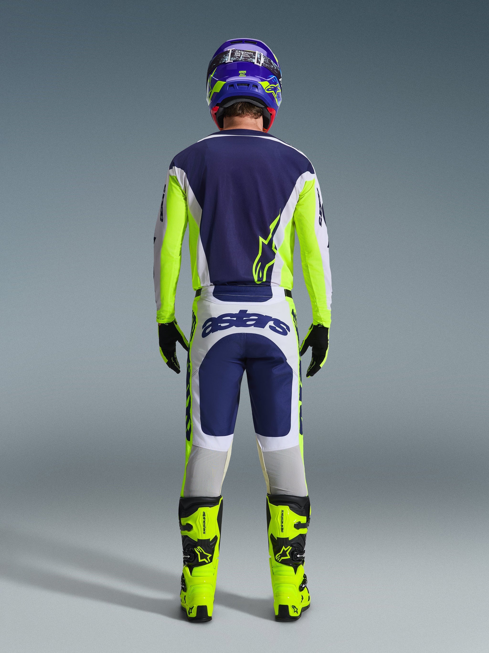 Vy bakifrån av en motocrossförare iklädd Alpinestars 2026 Racer Air Riway-tröja och matchande byxor i Light Gray Yellow Fluo Blue, kombinerat med neongula stövlar och en matchande hjälm, som visar en komplett uppsättning racingutrustning för offroad.