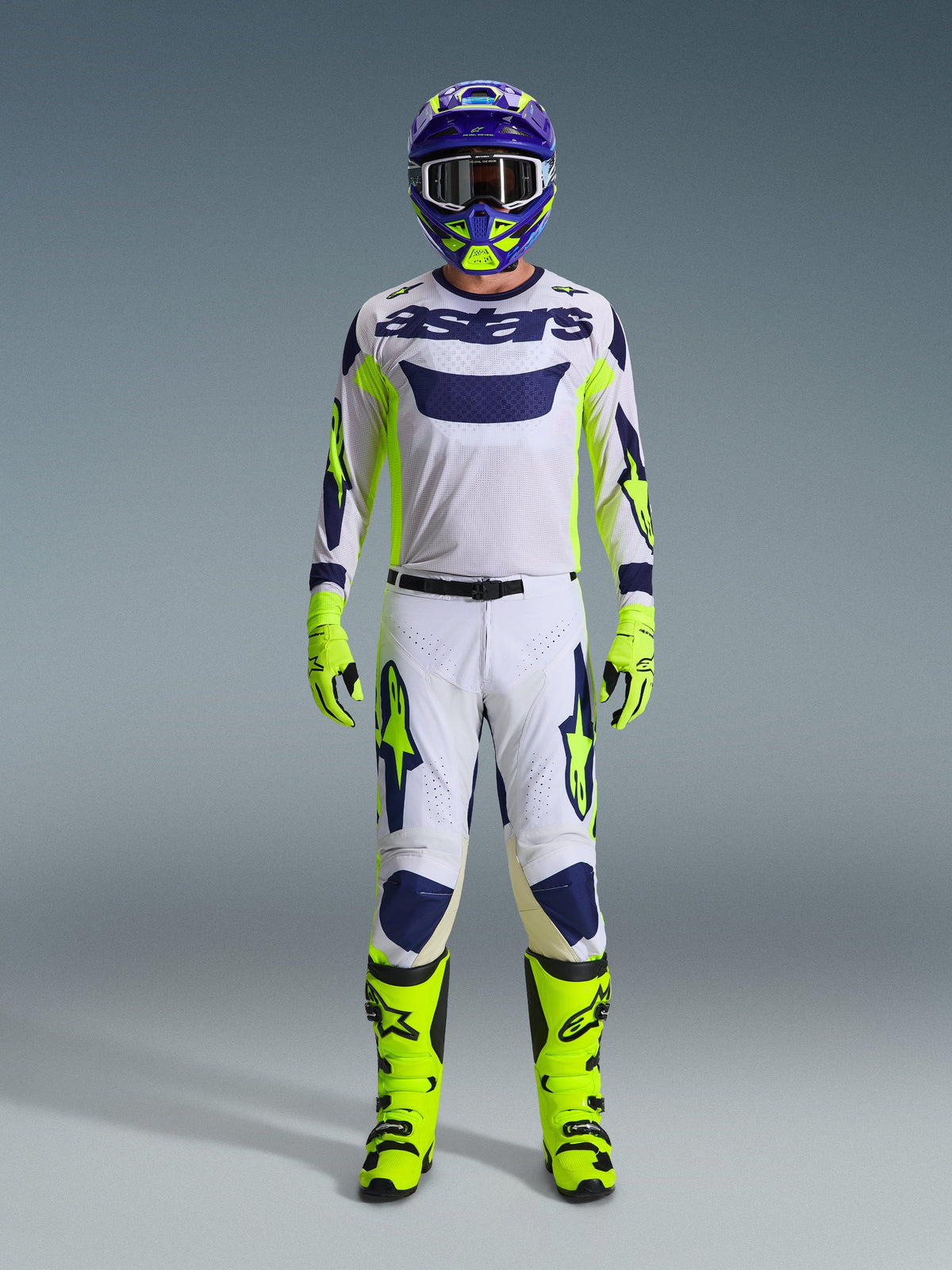 Motocrossförare i komplett racingutrustning, ljusgrå Racer Air Riway-tröja med neongula och blå detaljer, matchande vita och gula byxor, neongula stövlar och handskar, lila och gul hjälm, professionell utrustning för terrängkörning
