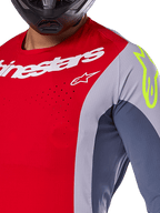Alpinestars 2025 Techstar Melt Jersey, motocrosströja, klar röd och grå med mörkblå paneler, med vit Alpinestars-logotyp och en neongul logotyp på axeln, närbild som visar laserskurna ventilationsperforeringar och en svart hjälm.