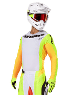 Motocrossförare klädd i Alpinestars 2025 Techstar Melt-utrustningsset, vit tröja med färgstarka gula och orangea ärmar, svart Alpinestars-logotyp på bröstet, matchande neongula handskar, vit hjälm med rödfärgade goggles, professionellt racingställ för terrängkörning