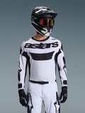 Motocrossförare i Alpinestars 2026 Racer Riway-tröja i vitt och svart, med 'astars'-logotyp på bröstet, matchande vita och svarta byxor, svart hjälm med goggles och svarta handskar, koordinerat ställ för terrängkörning