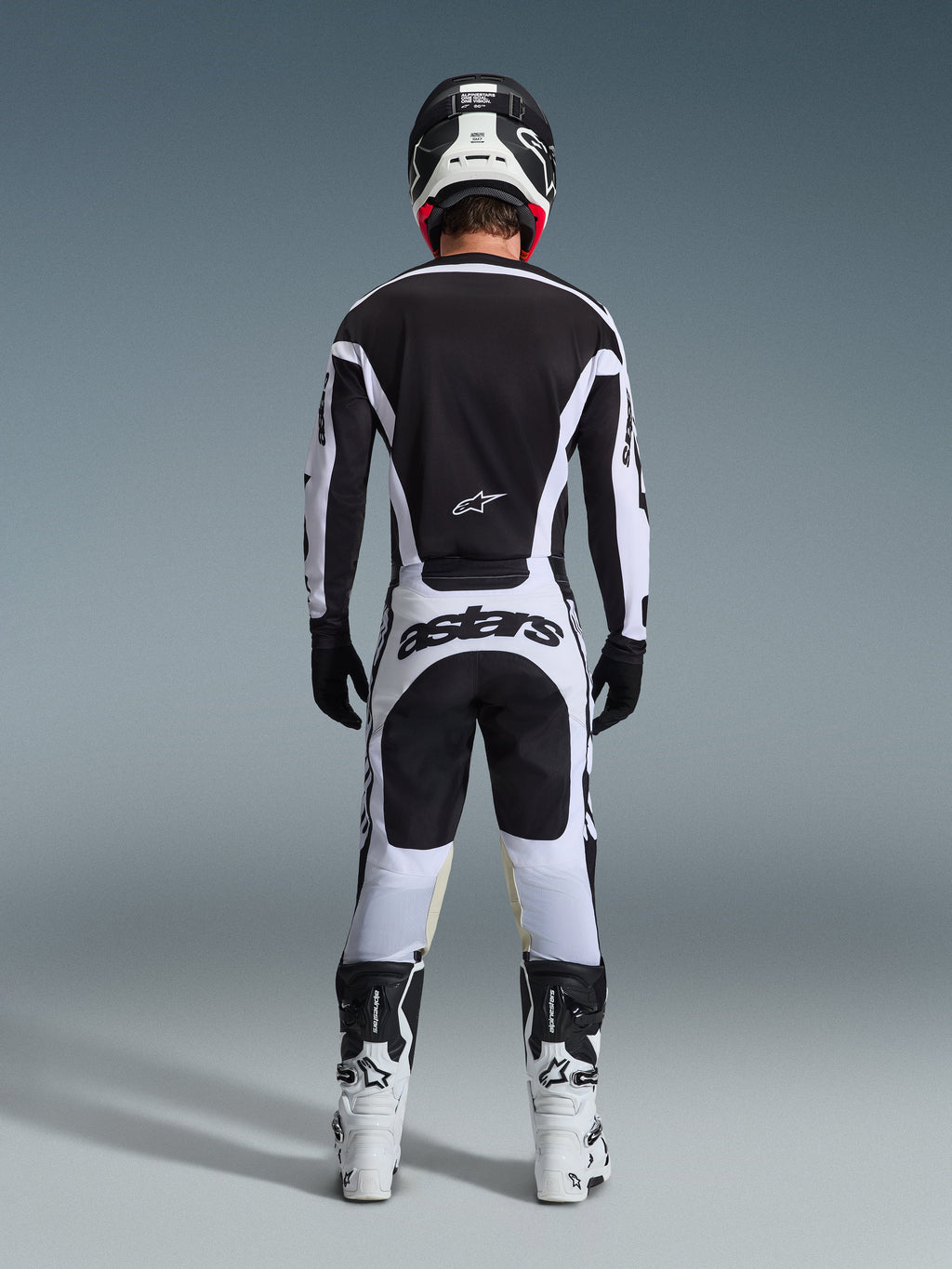 Motocrossförare sedd bakifrån i komplett racingutrustning, 2026 Racer Riway-tröja i vitt och svart, matchande byxor med 'astars'-logotyp på sätet, svart hjälm med vita detaljer, svarta handskar och vita och svarta stövlar, koordinerad professionell outfit för terrängkörning