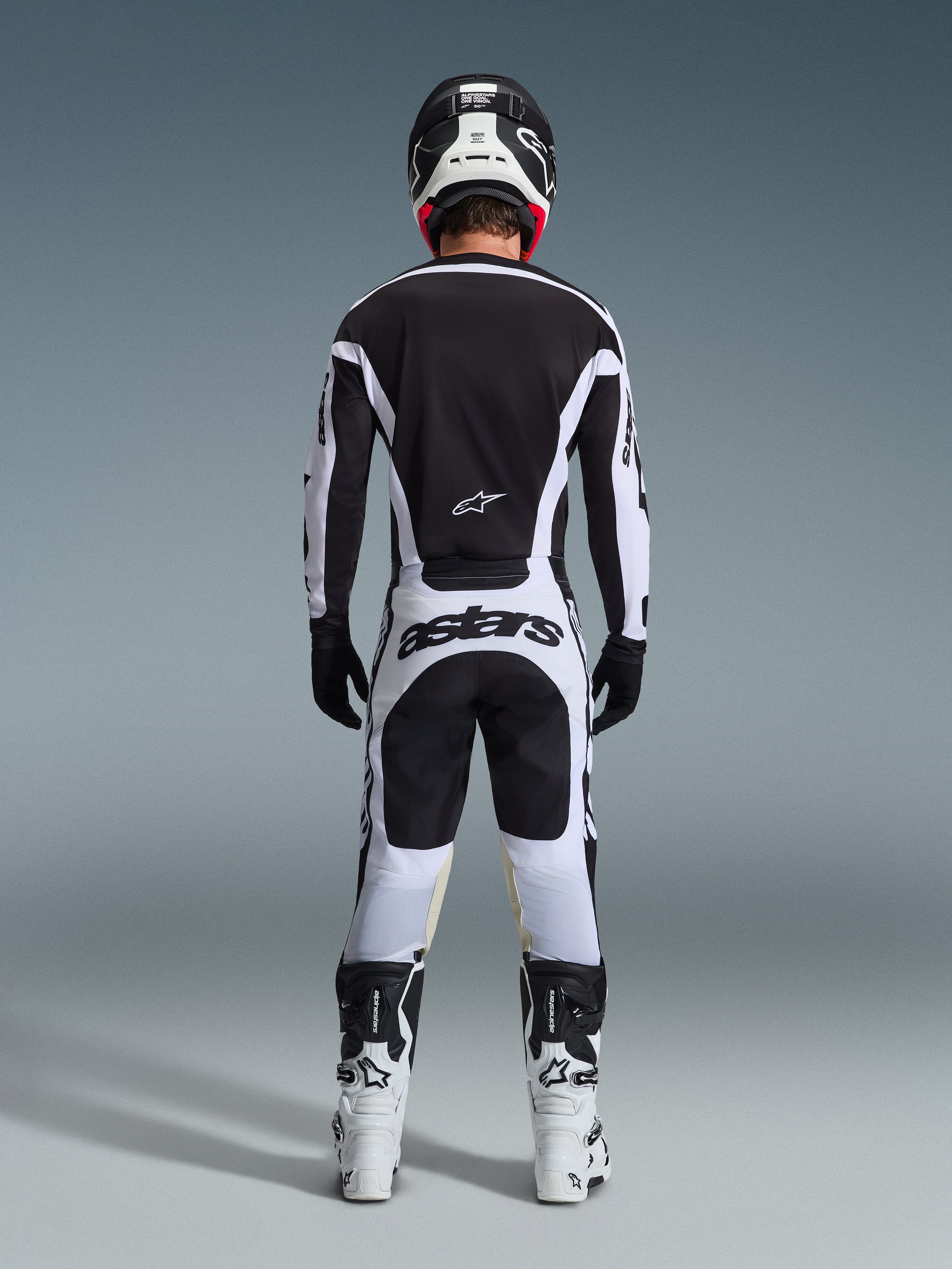 Motocrossförare sedd bakifrån i komplett racingutrustning, 2026 Racer Riway-tröja i vitt och svart, matchande byxor med 'astars'-logotyp på sätet, svart hjälm med vita detaljer, svarta handskar och vita och svarta stövlar, koordinerad professionell outfit för terrängkörning
