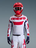 Motocrossförare iförd Alpinestars 2026 Racer Riway-tröja i vitt, blått och rött, med en stor röd 'astars'-logotyp på bröstet, marinblå sidopaneler, matchande vita och röda motocrossbyxor, röd hjälm med vita goggles och marinblå handskar, komplett utrustningsset för terrängkörning.