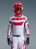 Motocrossförare iförd Alpinestars 2026 Racer Riway-tröja i vitt, blått och rött, med en stor röd 'astars'-logotyp på bröstet, marinblå sidopaneler, matchande vita och röda motocrossbyxor, röd hjälm med vita goggles och marinblå handskar, komplett utrustningsset för terrängkörning.