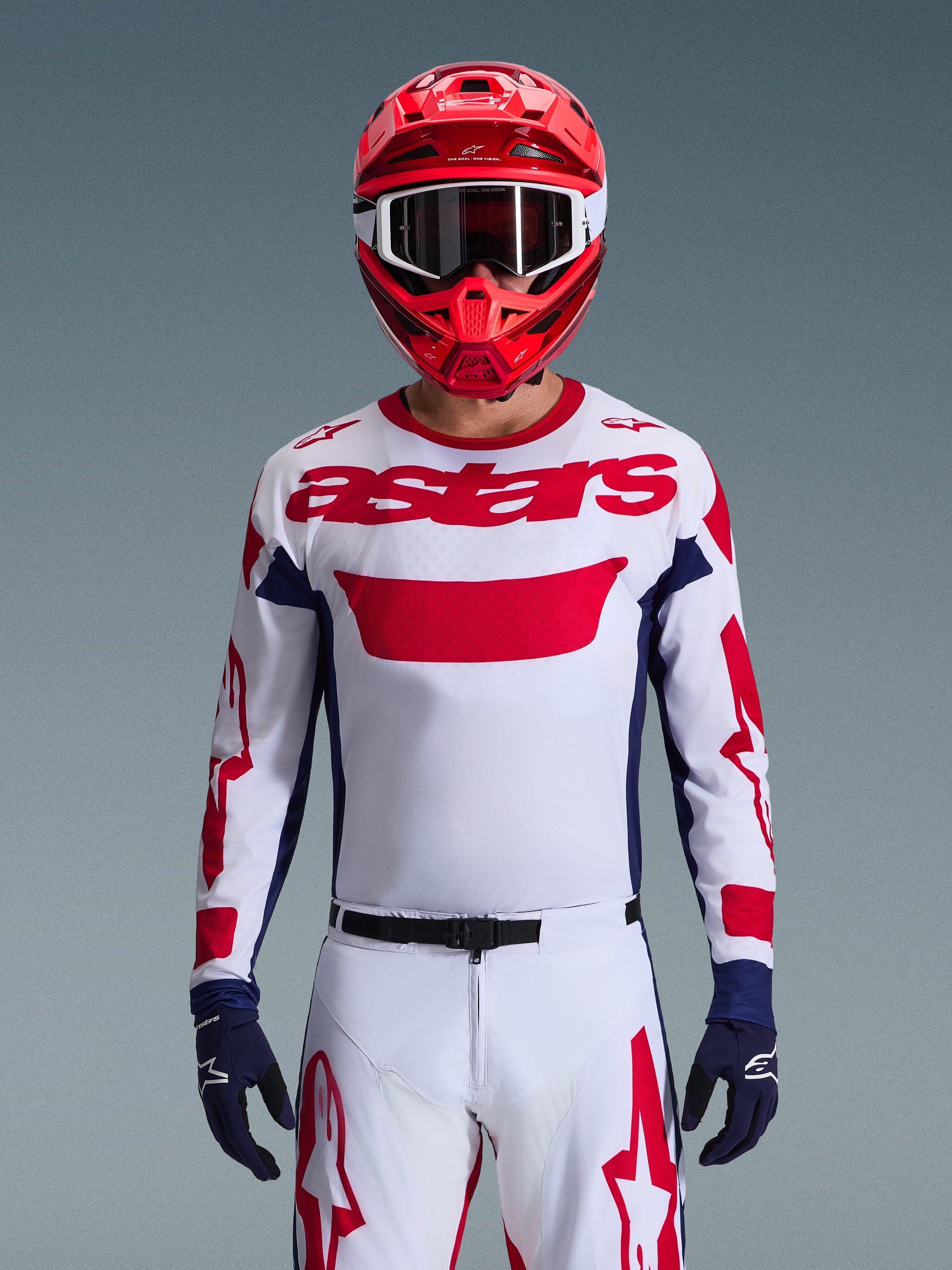 Motocrossförare iförd Alpinestars 2026 Racer Riway-tröja i vitt, blått och rött, med en stor röd 'astars'-logotyp på bröstet, marinblå sidopaneler, matchande vita och röda motocrossbyxor, röd hjälm med vita goggles och marinblå handskar, komplett utrustningsset för terrängkörning.