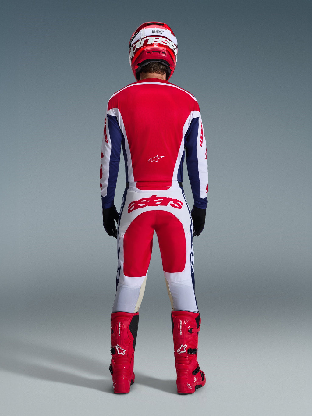 Motocrossförare sedd bakifrån i komplett racingutrustning, Alpinestars 2026 Racer Riway-tröja i White Blue Red, matchande vita och röda byxor med framträdande "astars"-logotyp, röda stövlar och röd och vit hjälm, koordinerad outfit för terrängkörning.