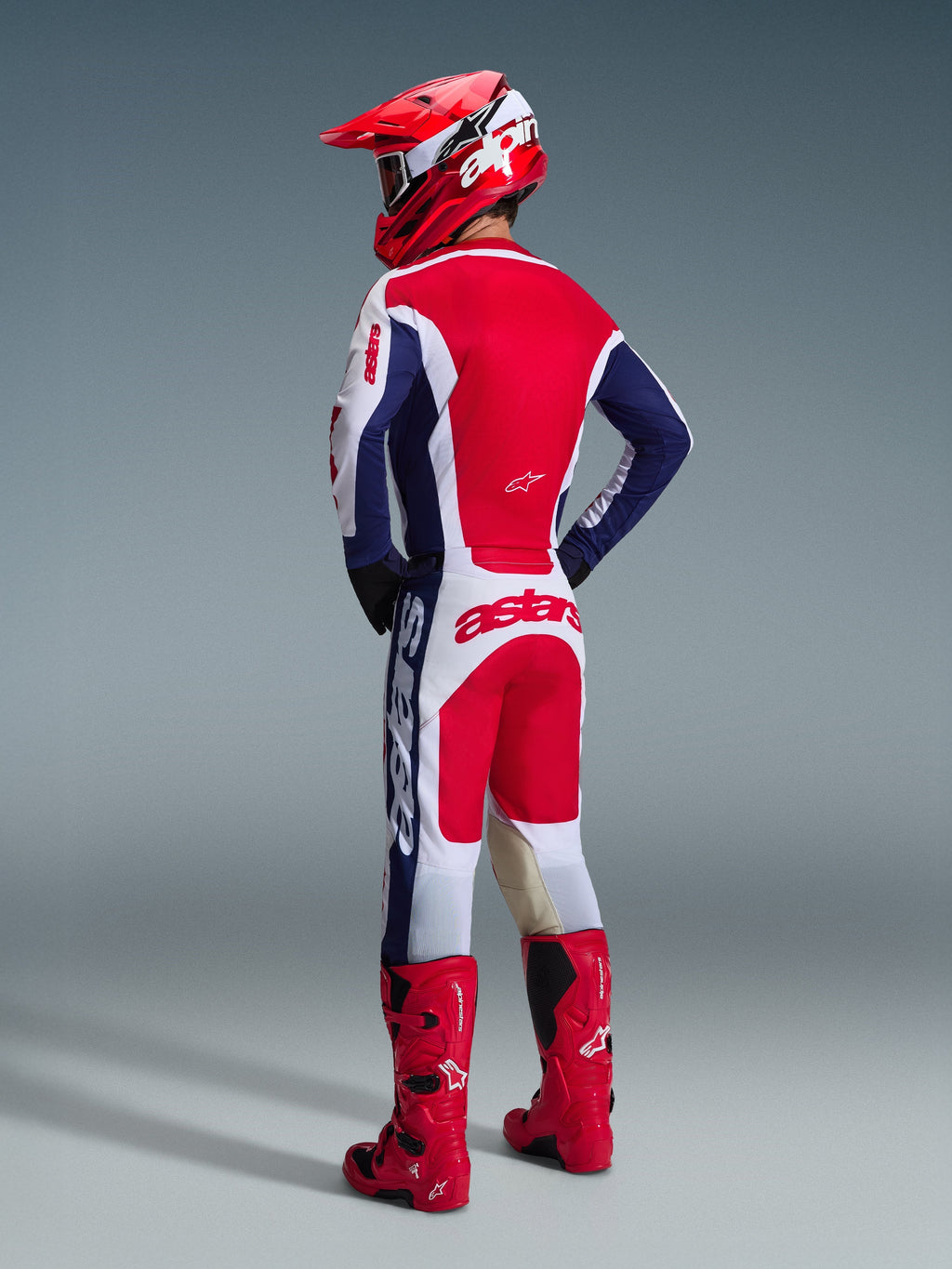 Motocrossförare sedd bakifrån iklädd Alpinestars 2026 Racer Riway-utrustningsset i vitt, blått och rött, inklusive en långärmad tröja med Astars-logotyp och matchande byxor, koordinerat med en röd hjälm och röda offroad-stövlar.