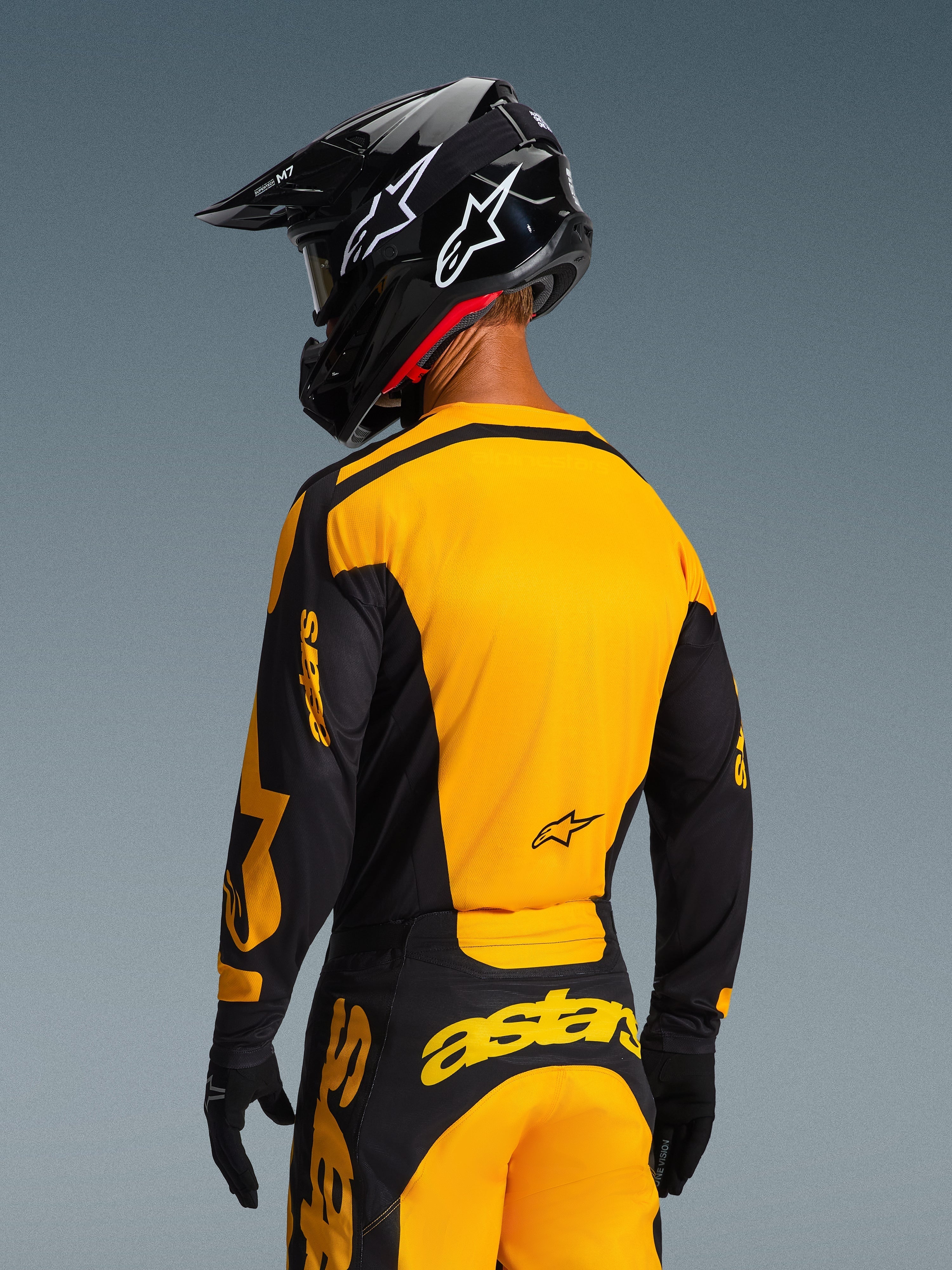 Motocrossförare sedd bakifrån i 2026 Racer Riway-tröja och matchande byxor i gult och svart, med framträdande Alpinestars-logotyper, svart hjälm med vita stjärnlogotyper och svarta handskar, koordinerat motocross-utrustningsset för terrängkörning.