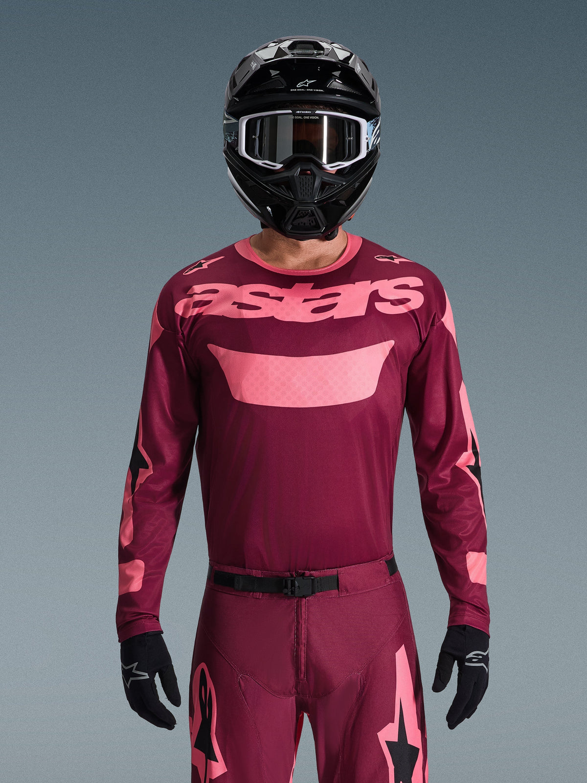 Motocrossförare klädd i Alpinestars 2026 Racer Riway-tröja i Dark Red Coral, med en vinröd bas och korallrosa 'astars'-logotyp på bröstet, matchad med tillhörande motocrossbyxor, svart hjälm och svarta handskar, professionellt utrustningsset för terrängkörning.