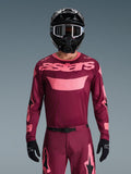 Motocrossförare klädd i Alpinestars 2026 Racer Riway-tröja i Dark Red Coral, med en vinröd bas och korallrosa 'astars'-logotyp på bröstet, matchad med tillhörande motocrossbyxor, svart hjälm och svarta handskar, professionellt utrustningsset för terrängkörning.