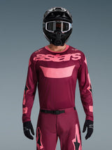 Motocross rider wearing the Alpinestars 2026 Racer Riway Jersey in Dark Red Coral, featuring a maroon body with coral pink 'astars' chest branding, paired with matching motocross pants, black helmet, and black gloves, professional gear set for off-road racing.