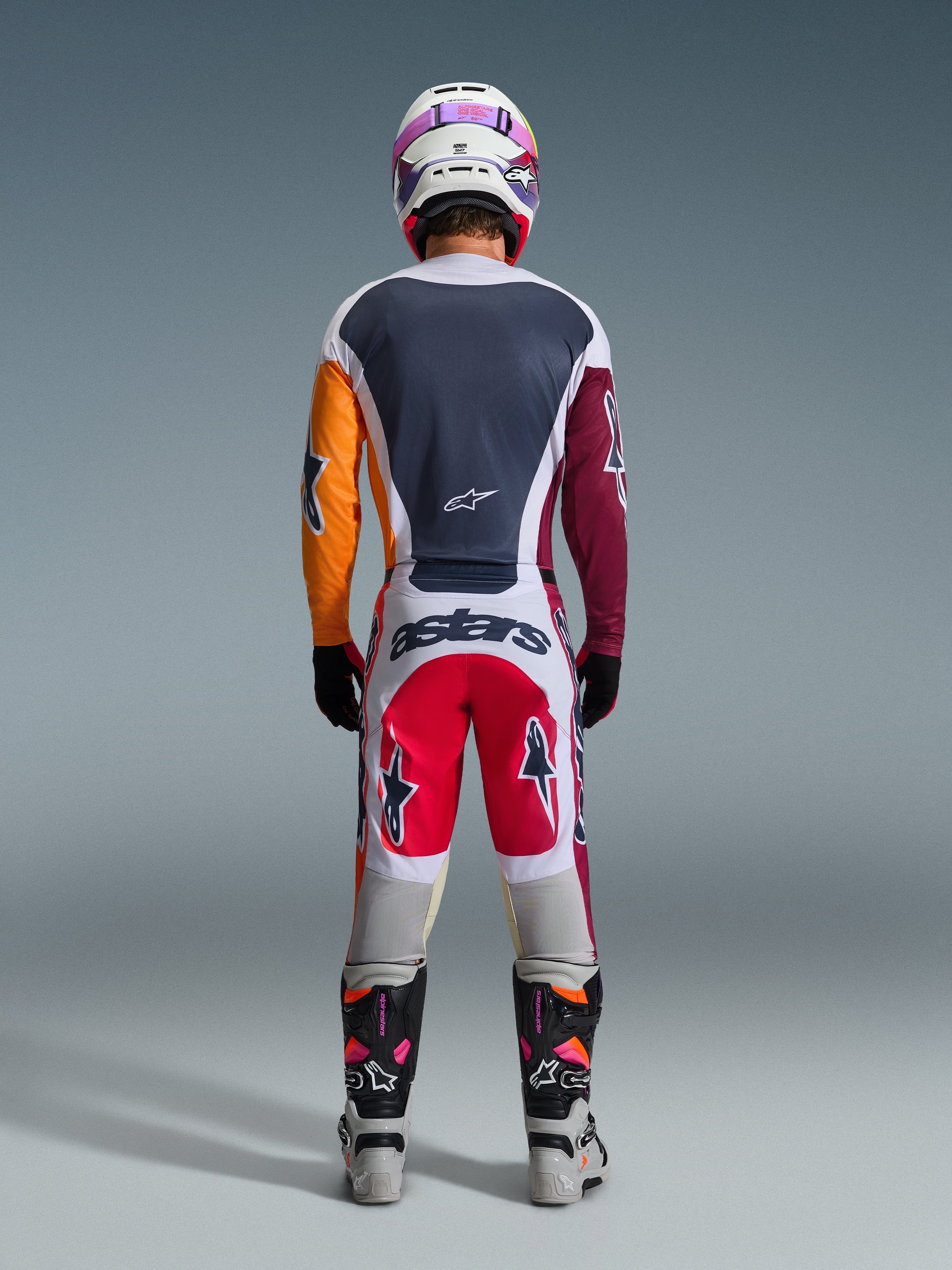 Motocrossförare sedd bakifrån i komplett racingutrustning, 2026 Racer Portl-tröja i rött, ljusgrått och mörkgrönt med asymmetriska orangea och vinröda ärmar, matchande vita och röda byxor, vit hjälm med lila goggles, vita och svarta stövlar, koordinerad outfit för terrängkörning