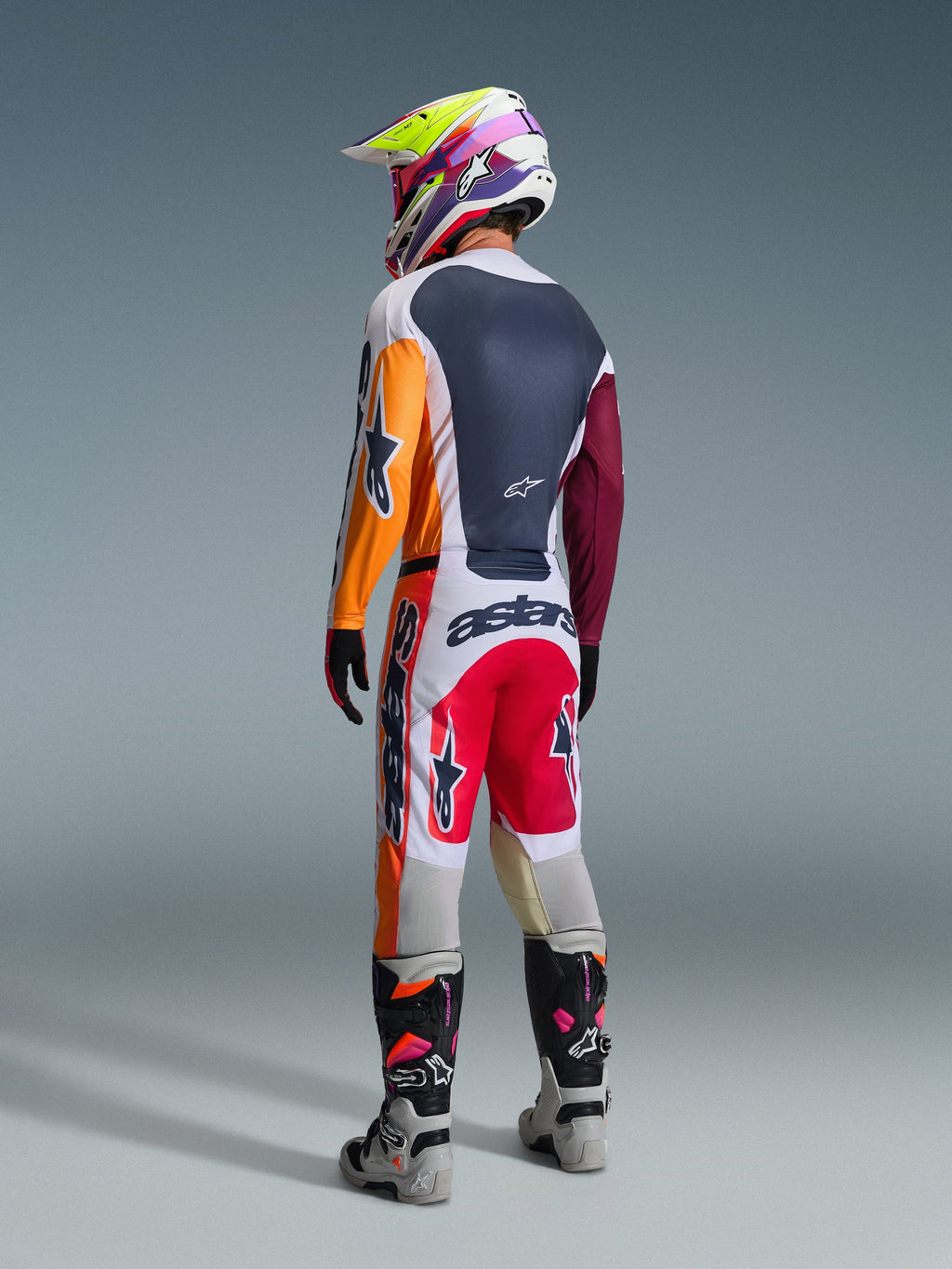 Motocrossförare i komplett racingutrustning sedd bakifrån, Alpinestars 2026 Racer Portl-tröja i Red Light Gray och Dark Green med orangea och vinröda paneler, matchande vita och röda byxor, grå och svarta stövlar med rosa detaljer, flerfärgad hjälm, koordinerad outfit för terrängkörning.