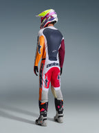Motocrossförare i komplett racingutrustning sedd bakifrån, Alpinestars 2026 Racer Portl-tröja i Red Light Gray och Dark Green med orangea och vinröda paneler, matchande vita och röda byxor, grå och svarta stövlar med rosa detaljer, flerfärgad hjälm, koordinerad outfit för terrängkörning.