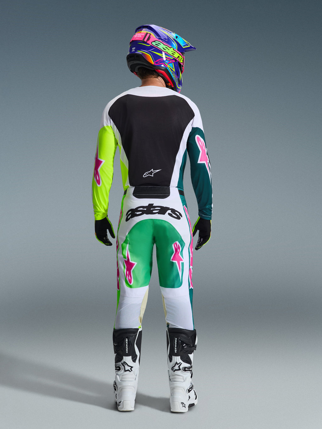 Rear view of motocross rider wearing the Alpinestars 2026 Racer Portl Gear Set, white jersey with a black back panel and neon yellow and dark green sleeves featuring pink stars, matching white pants with green and teal accents and pink star logos, coordinated with a colorful helmet, black gloves, and white and black boots.