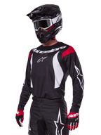 Motocrossförare i Alpinestars 2025 Fluid Haul utrustningsset, svart tröja med röda axelpaneler och vit grafik, matchande svarta byxor med vita stjärnlogotyper, röda och svarta handskar, svart hjälm med vita detaljer och goggles, koordinerad motocrossutrustning