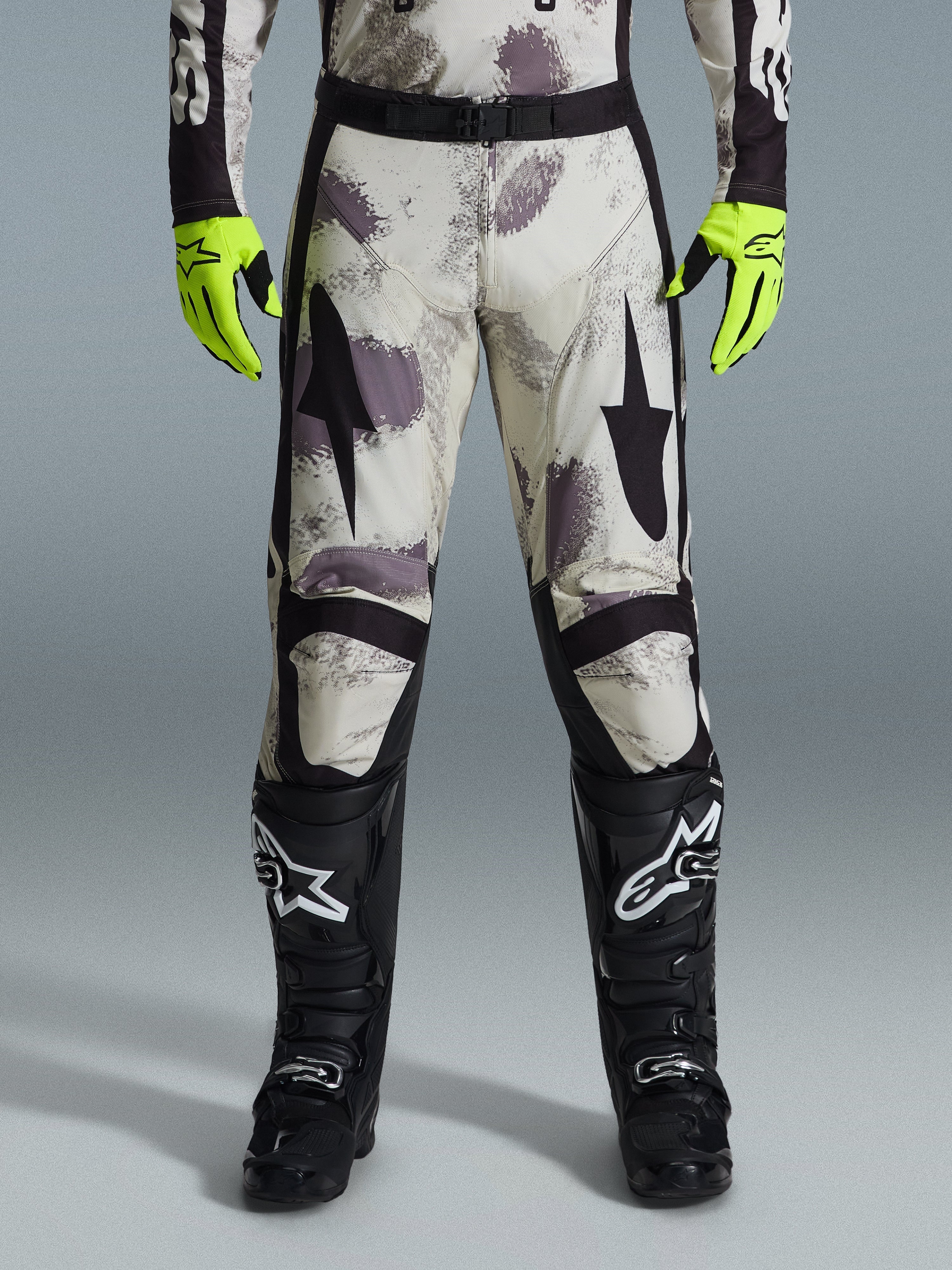 Alpinestars 2026 Racer Lahnd byxor, motocrossbyxor, Sand Camo-mönster med grå och lila abstrakt grafik, bärs med neongula handskar och svarta stövlar, svarta stjärnlogotyper på låren och ett svart midjebälte, hållbar racingutrustning för terrängkörning