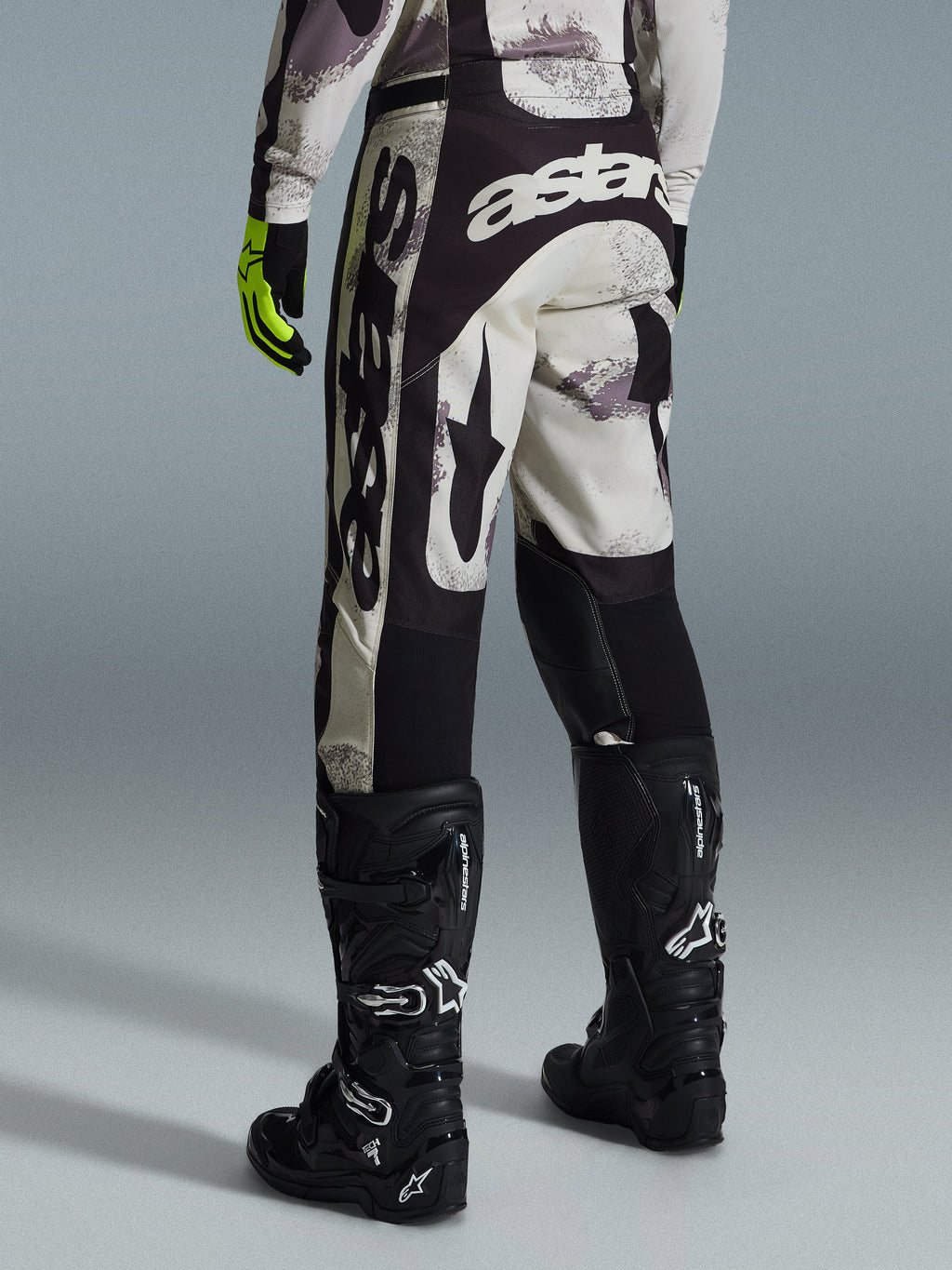 Alpinestars 2026 Racer Lahnd byxor, motocrossbyxor, sand-camo och grå, vy bakifrån med tydlig vit logotyp på sätet och benen, kombinerade med svarta motocross-stövlar och gula handskar, professionell racingutrustning för terrängkörning
