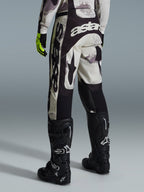 Alpinestars 2026 Racer Lahnd byxor, motocrossbyxor, sand-camo och grå, vy bakifrån med tydlig vit logotyp på sätet och benen, kombinerade med svarta motocross-stövlar och gula handskar, professionell racingutrustning för terrängkörning