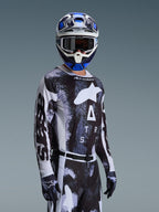 Motocrossförare i Alpinestars 2026 Racer Lahnd-tröja i Iron Camo, med ett svart och grått kamouflagemönster och en stor vit Alpinestars-logotyp på bröstet, matchad med hjälm och handskar för terrängkörning.