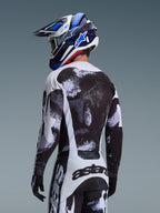 Alpinestars 2026 Racer Lahnd Jersey i Iron Camo, vy bakifrån på en motocrossförare i ett svart, vitt och grått abstrakt mönstrat ställ, inklusive matchande byxor, svarta handskar och en blå racinghjälm med goggles.