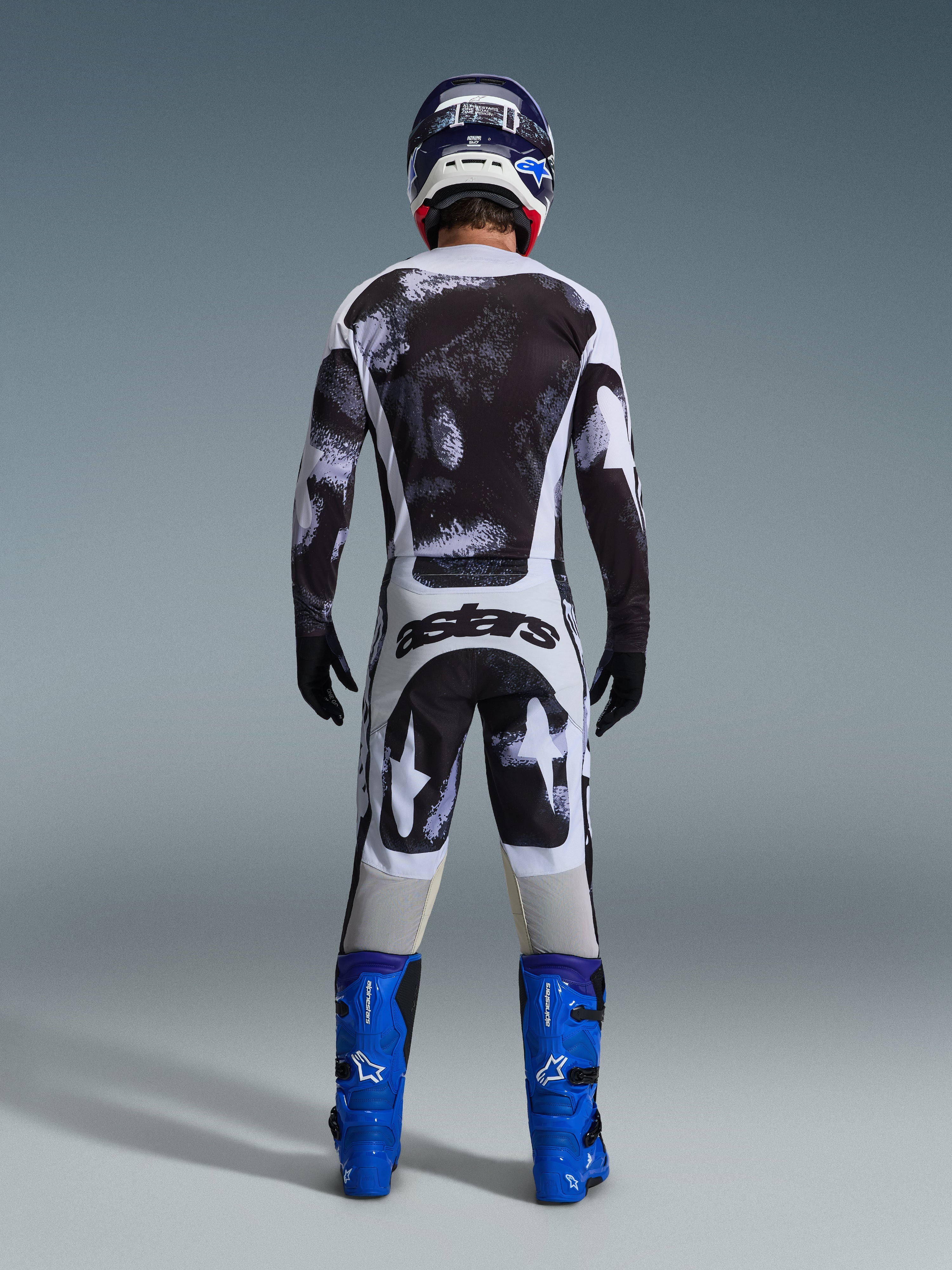 Motocross rider from the rear wearing the Alpinestars 2026 Racer Lahnd Gear Set, black and white abstract patterned jersey and pants, 'astars' branding on the seat, paired with blue boots and a matching helmet, professional off-road racing apparel.